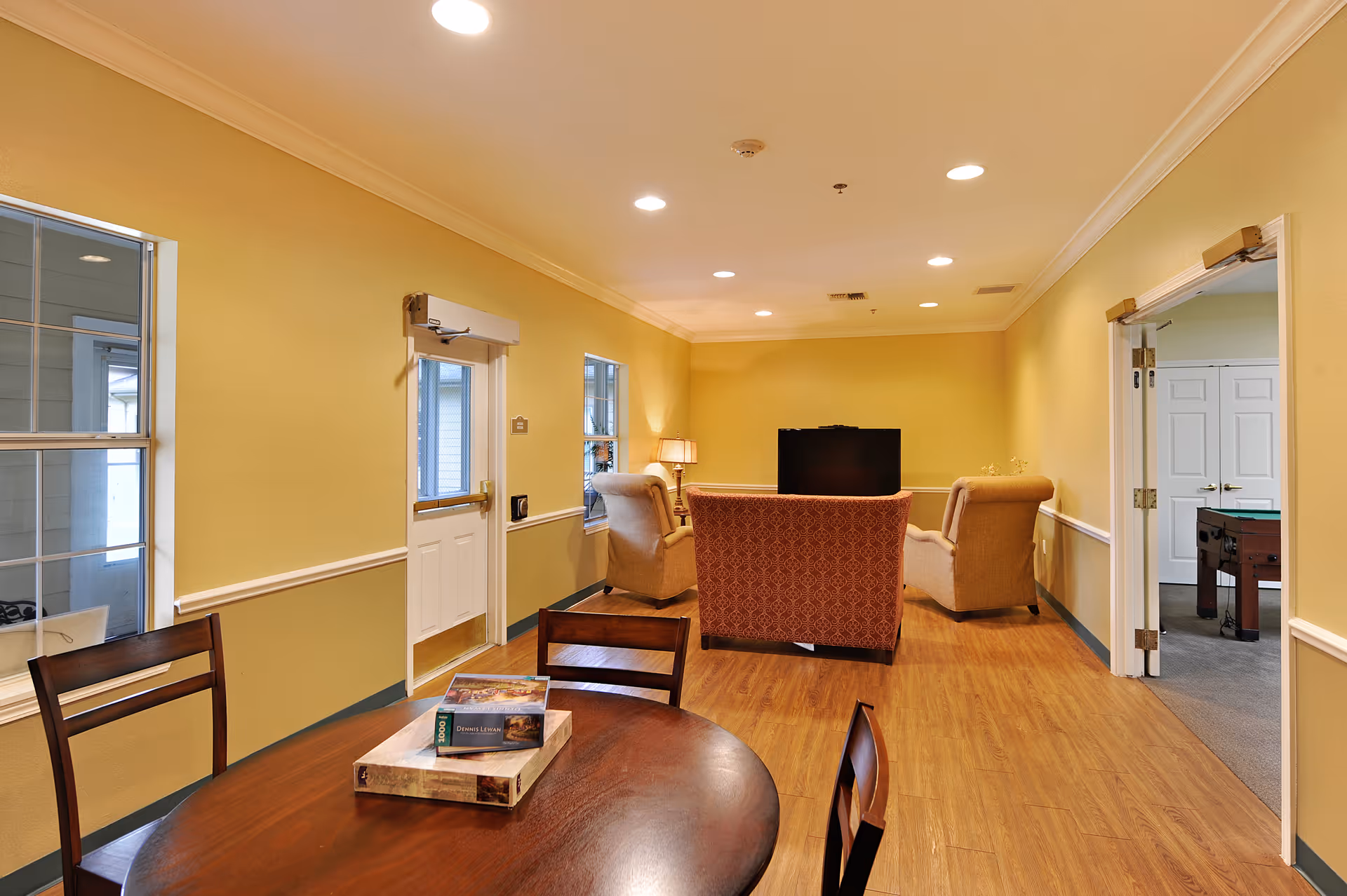 Community lounge with a round table in the foreground and armchairs facing a television in a warmly lit room.