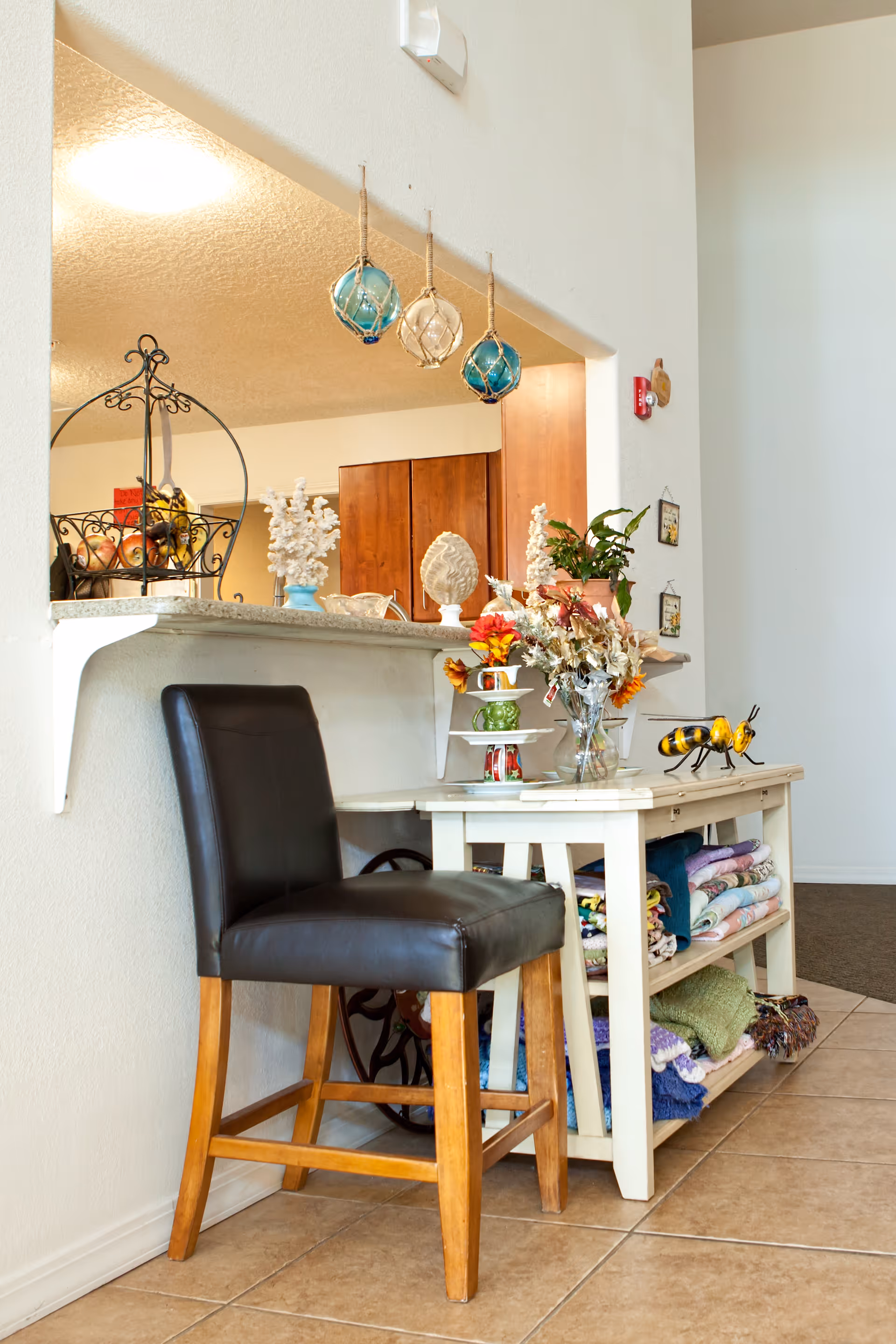 Interior view of a cozy corner in an assisted living facility featuring a black leather chair with wooden legs next to a white table holding a vase with flowers, decorative items, and neatly folded blankets on the lower shelves. Above the table, three hanging glass buoy decorations are suspended from the ceiling. In the background, a kitchen area with wooden cabinets is partially visible.