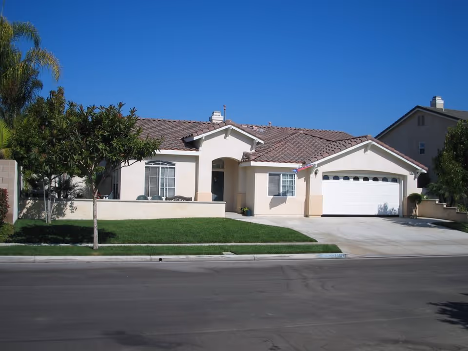 Front exterior of a single-story house with a tiled roof, attached two-car garage, small lawn and a tree under a clear blue sky.