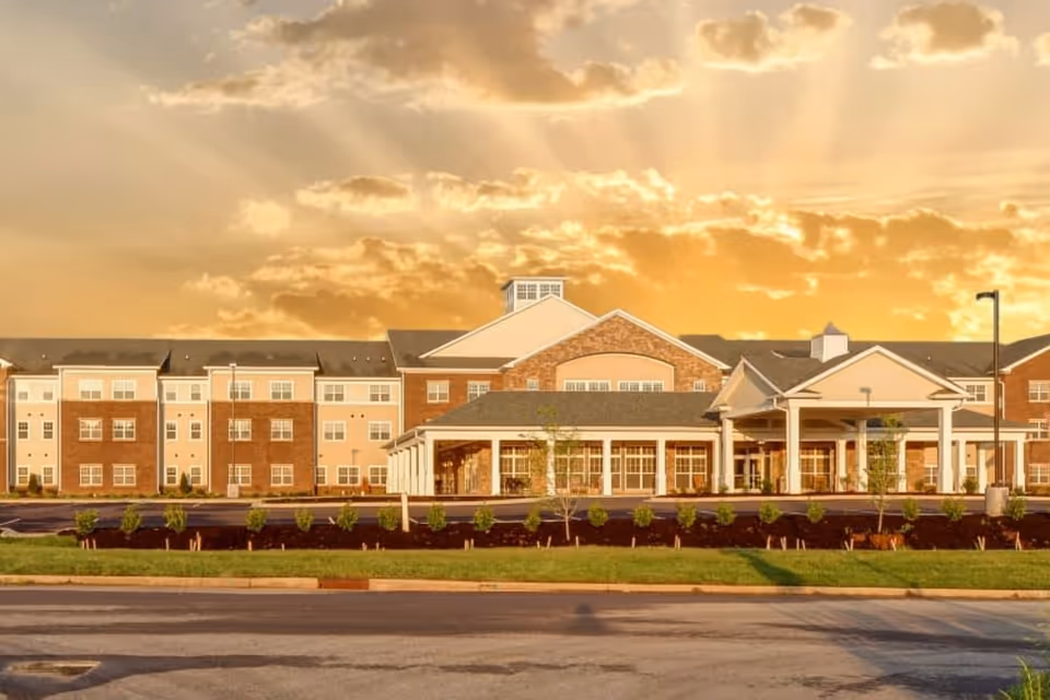 Exterior view of a large senior living facility building with a brick and beige facade under a dramatic sky with sun rays breaking through clouds at sunset.