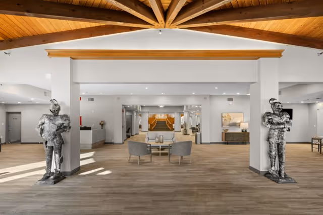 Spacious senior living community interior with wooden vaulted ceiling beams, two medieval knight statues on either side of the entrance, a seating area with two chairs and a small table in the center, and a staircase in the background.