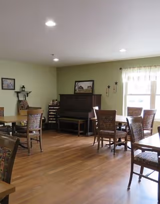 A well-lit room with wooden flooring featuring several wooden tables and chairs arranged for seating. There is a piano against the far wall with a framed picture on top, and a window with sheer curtains allowing natural light to enter. The walls are painted light green, and there are some decorative items on the walls and shelves.