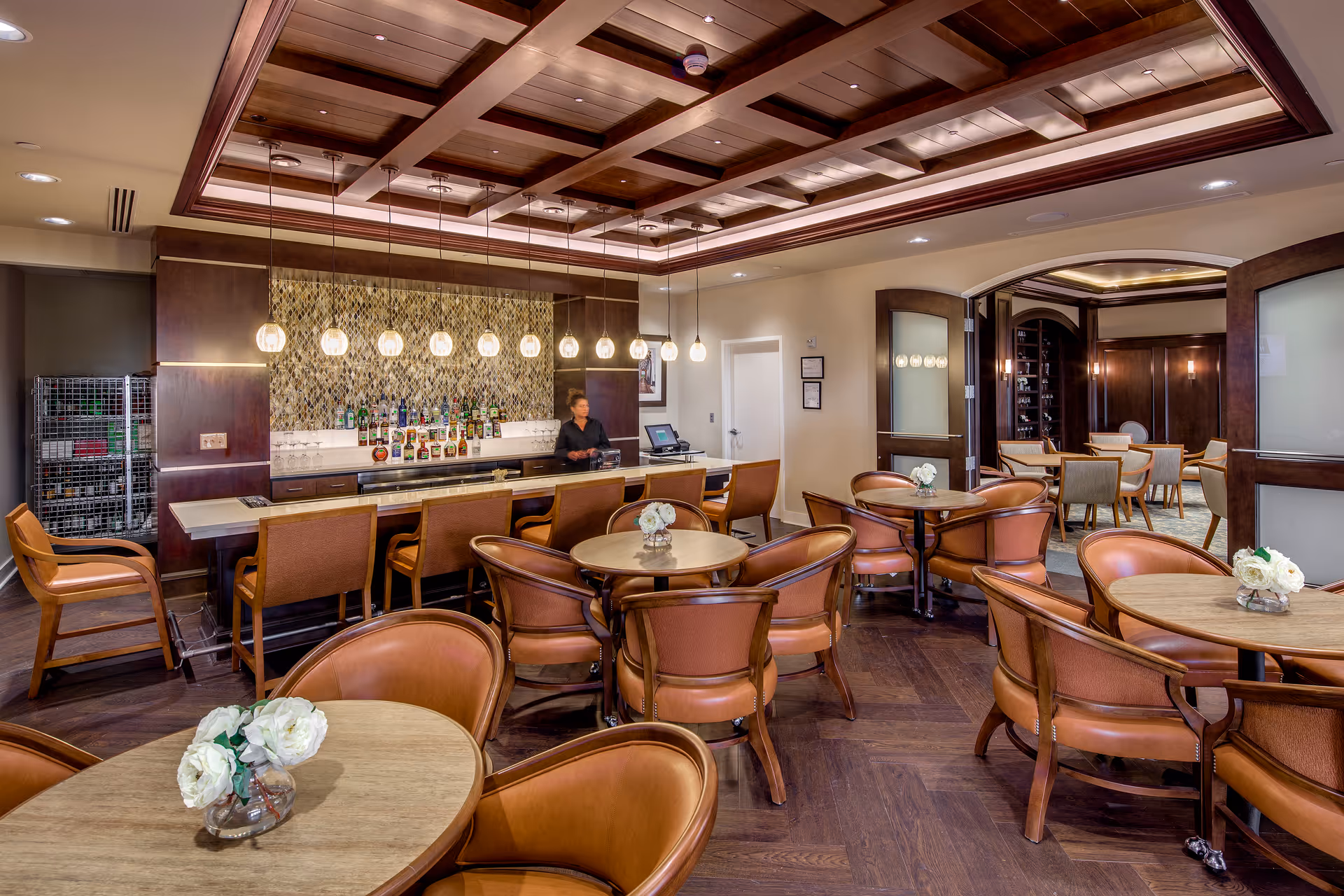 A warm and inviting lounge area with round tables and brown leather chairs arranged neatly. In the background, there is a bar with a variety of bottles displayed on shelves, illuminated by hanging pendant lights. A staff member stands behind the bar. The room features a coffered ceiling with recessed lighting and dark wood flooring. Adjacent to the bar area, there is another seating area visible through open double doors.