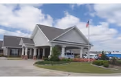 Exterior view of a single-story nursing and rehab facility building with a covered entrance, an American flag on a flagpole, and a driveway with parked vehicles under a partly cloudy sky.