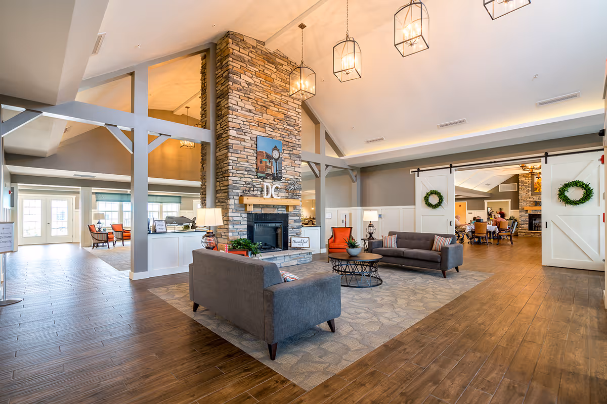 Spacious senior living community lounge with seating around a stone fireplace, high ceilings, and pendant lights.