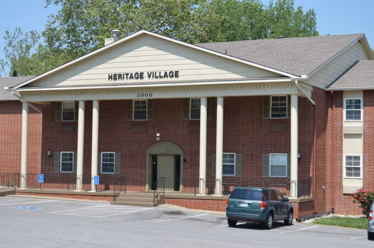 Exterior front view of a two-story brick building with white columns and a beige triangular pediment displaying the name 'HERITAGE VILLAGE'. There are several windows with green shutters, a central entrance with steps and a ramp, and a parking lot with a green SUV parked in front.