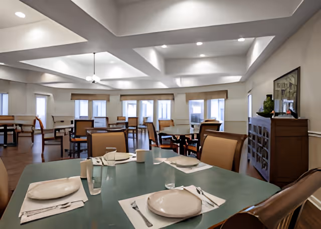 A spacious dining room with multiple tables and chairs arranged neatly. Each table is set with plates, napkins, utensils, and glasses. The room has large windows with blinds, a coffered ceiling with recessed lighting, and a sideboard with decorative items and a framed picture on the wall.