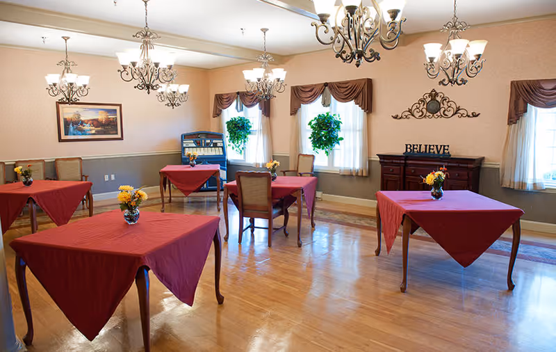 A dining room with wooden floors and several square tables covered with red tablecloths. Each table has a small vase with yellow flowers. The room has multiple chandeliers hanging from the ceiling, windows with brown valances and white curtains, and a sideboard with the word 'BELIEVE' displayed on it. There are also two green plants hanging in front of the windows and a framed picture on the wall.