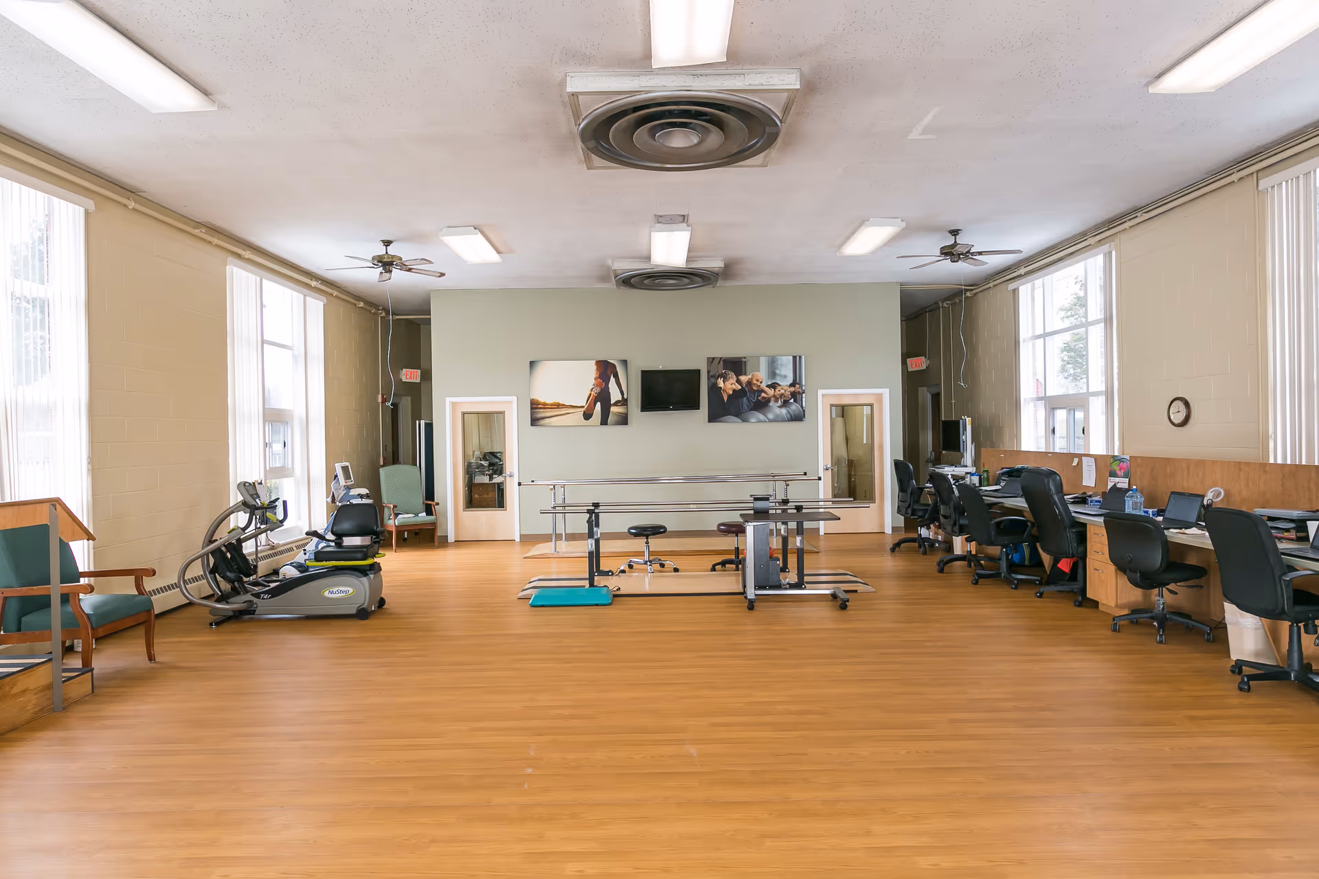Large rehabilitation room with exercise machines, parallel bars, therapy tables, desks and chairs under ceiling fans and fluorescent lights.