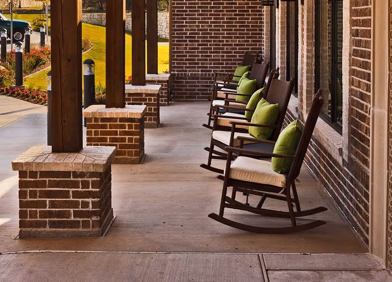 A row of wooden rocking chairs with green cushions lined up on a covered porch with brick pillars and walls, overlooking a landscaped outdoor area with grass and flowers.