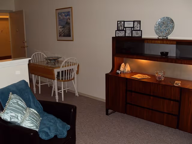 Interior view of a living area in an assisted living community featuring a dark armchair with blue cushions and a throw, a small wooden dining table with two white chairs, and a wooden cabinet with decorative items including framed photos, a mosaic plate, and small figurines.