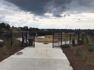 A paved walkway leading to a gated outdoor area with metal fencing on both sides. The sky is cloudy and overcast, and there are some small plants and shrubs along the sides of the walkway.