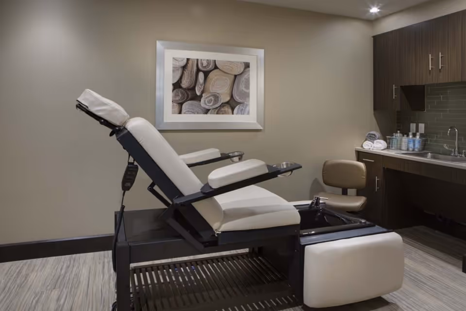 A modern pedicure spa chair with white cushions and a black foot basin in a well-lit room. The room has beige walls, a framed picture of smooth stones, wooden cabinets, a small beige chair, a countertop with rolled towels, and various spa supplies near a sink.
