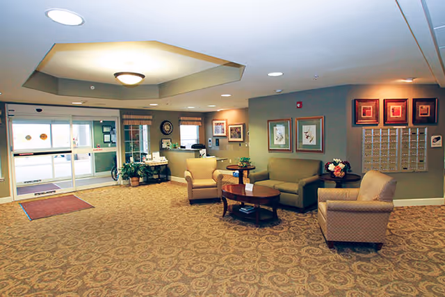 A spacious and well-lit lobby area with patterned carpet flooring, beige walls, and ceiling lights. The seating area includes a green sofa, two beige armchairs, and two wooden coffee tables with decorative items. On the walls are framed artwork and a set of mailboxes. The entrance features glass automatic sliding doors with a red mat in front.