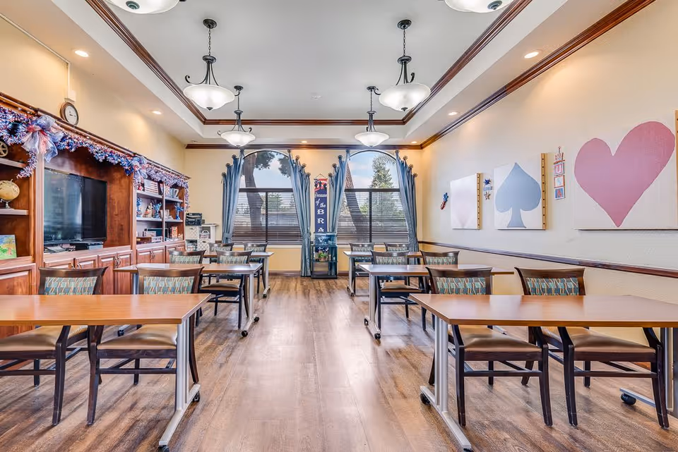 A bright and spacious room with multiple wooden tables and chairs arranged in rows. The room features large windows with blue curtains, decorative wall art with card suit symbols (diamond, spade, heart), and a wooden entertainment center with a television and shelves. The ceiling has elegant light fixtures and crown molding, and the floor is a wood finish.