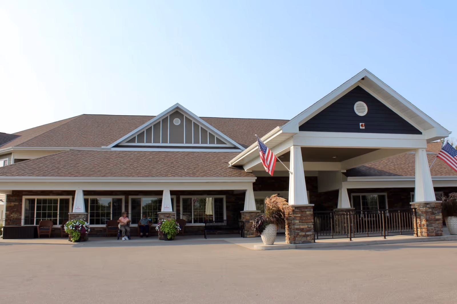 Front entrance of a single-story senior living facility with a covered porte-cochere, American flags, planters and benches with two people seated.