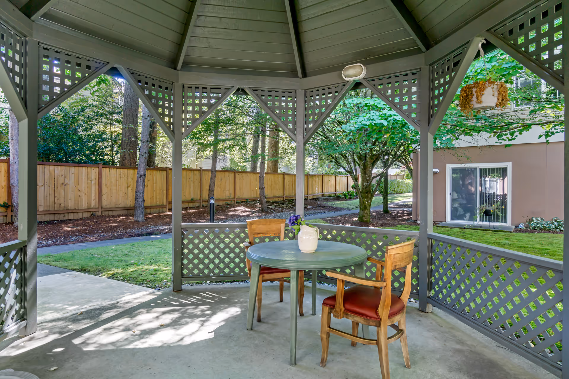 View from inside a gray wooden gazebo with lattice panels, containing a round table with a white pitcher and two wooden chairs with red cushions. The gazebo overlooks a green lawn, trees, a wooden fence, and a building with a sliding glass door.