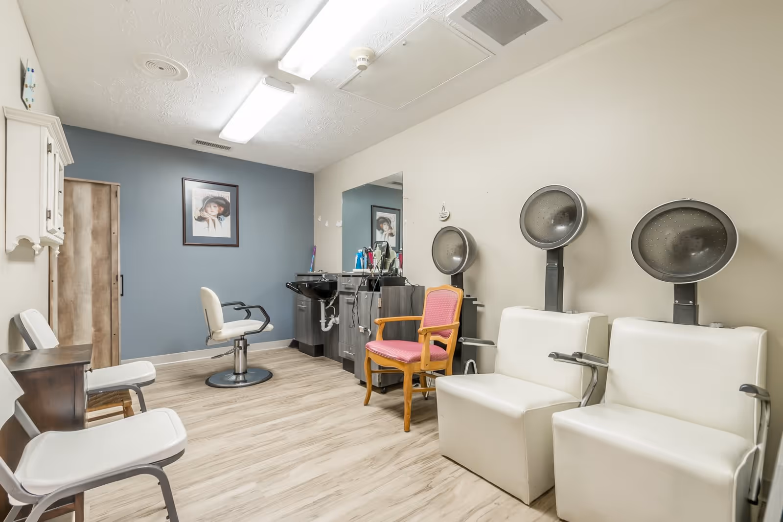 Interior of a hair salon area in a senior living facility with two white hair drying chairs, a pink cushioned wooden chair, a white salon chair in front of a sink, and a large mirror on the wall. The room has light-colored wood flooring, beige and blue walls, and a framed picture on the blue wall.