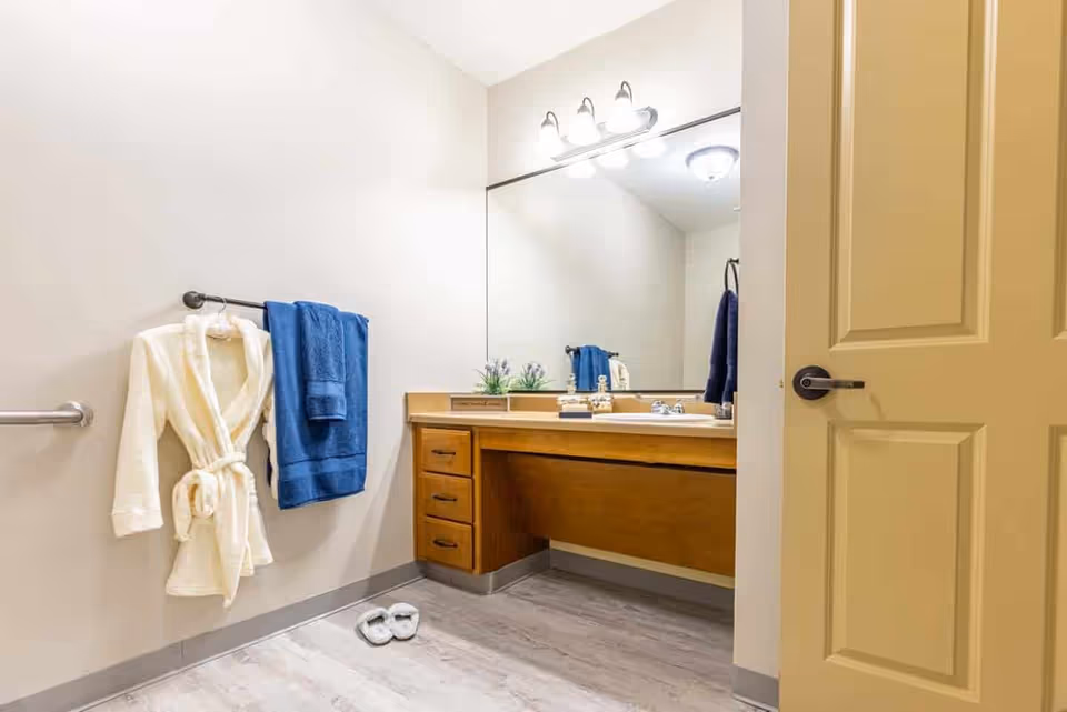 A clean bathroom with a large mirror above a wooden vanity with drawers. A white bathrobe and a blue towel hang on a towel rack on the wall. A pair of white slippers are placed on the floor. The bathroom has light-colored walls and flooring, and a partially open beige door is visible on the right.