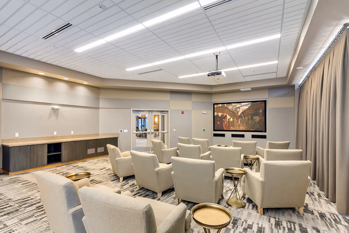 A modern media room with multiple beige armchairs arranged in rows facing a large wall-mounted screen displaying an image of an eagle. The room has a patterned carpet, a ceiling-mounted projector, and a long counter with cabinets along one wall. Large curtains cover the windows on the right side.