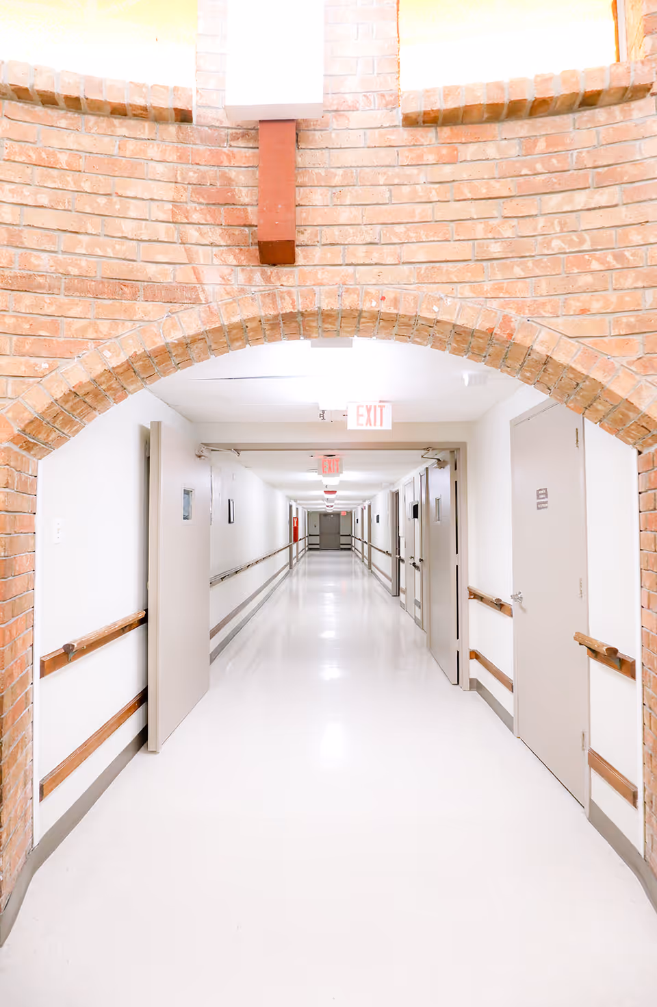 A long, well-lit hallway in a healthcare facility with white walls and floor, wooden handrails on both sides, multiple closed doors, and a brick archway at the entrance. Exit signs are visible on the ceiling.