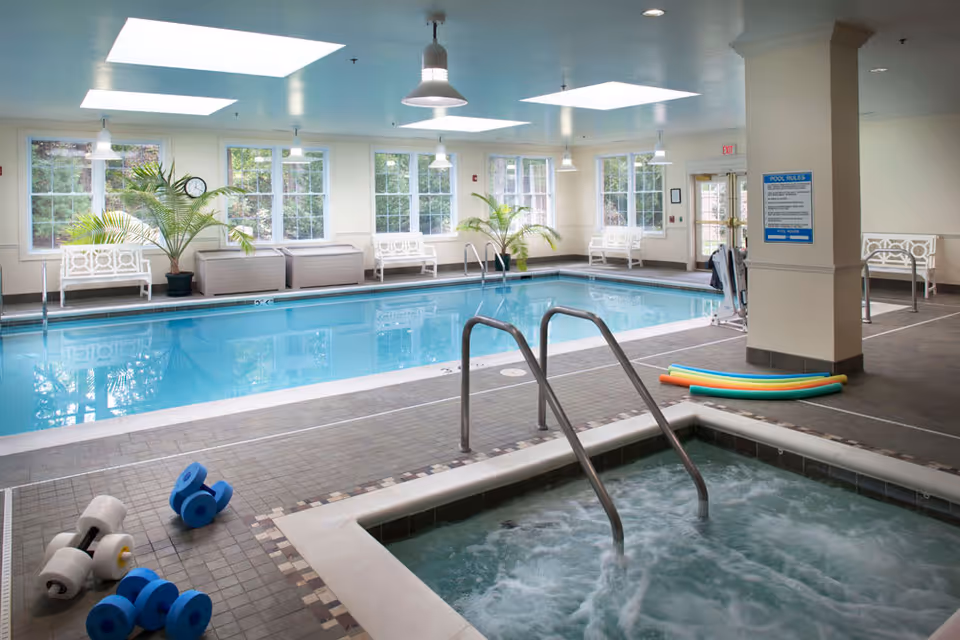 Indoor swimming pool area with a hot tub in the foreground, pool noodles on the floor, white benches along the windows, potted plants, and large skylights on the ceiling providing natural light.