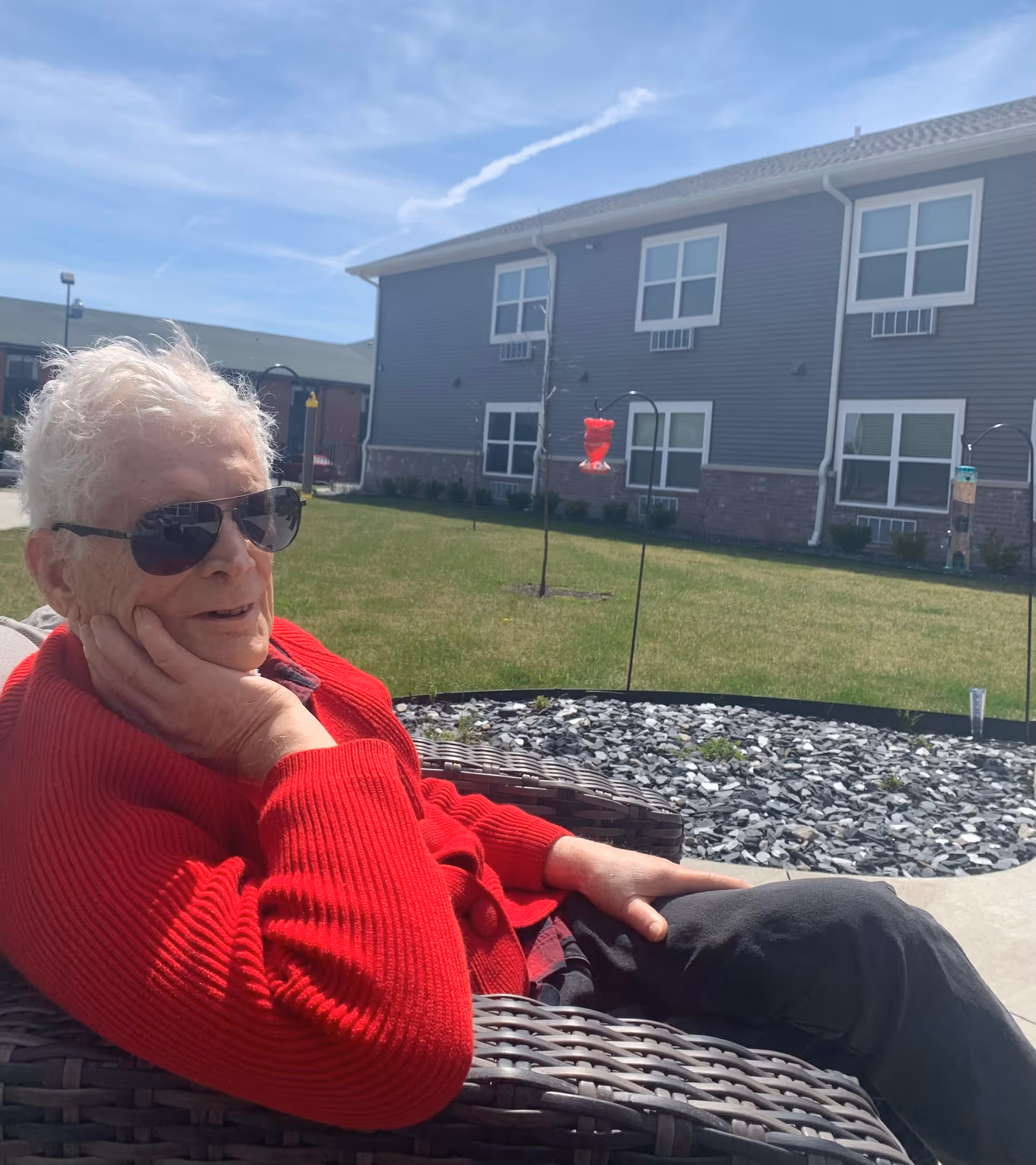 An elderly person wearing a red sweater and sunglasses is sitting comfortably in a wicker chair outdoors on a sunny day. Behind them is a grassy area with bird feeders and a two-story building with multiple windows.