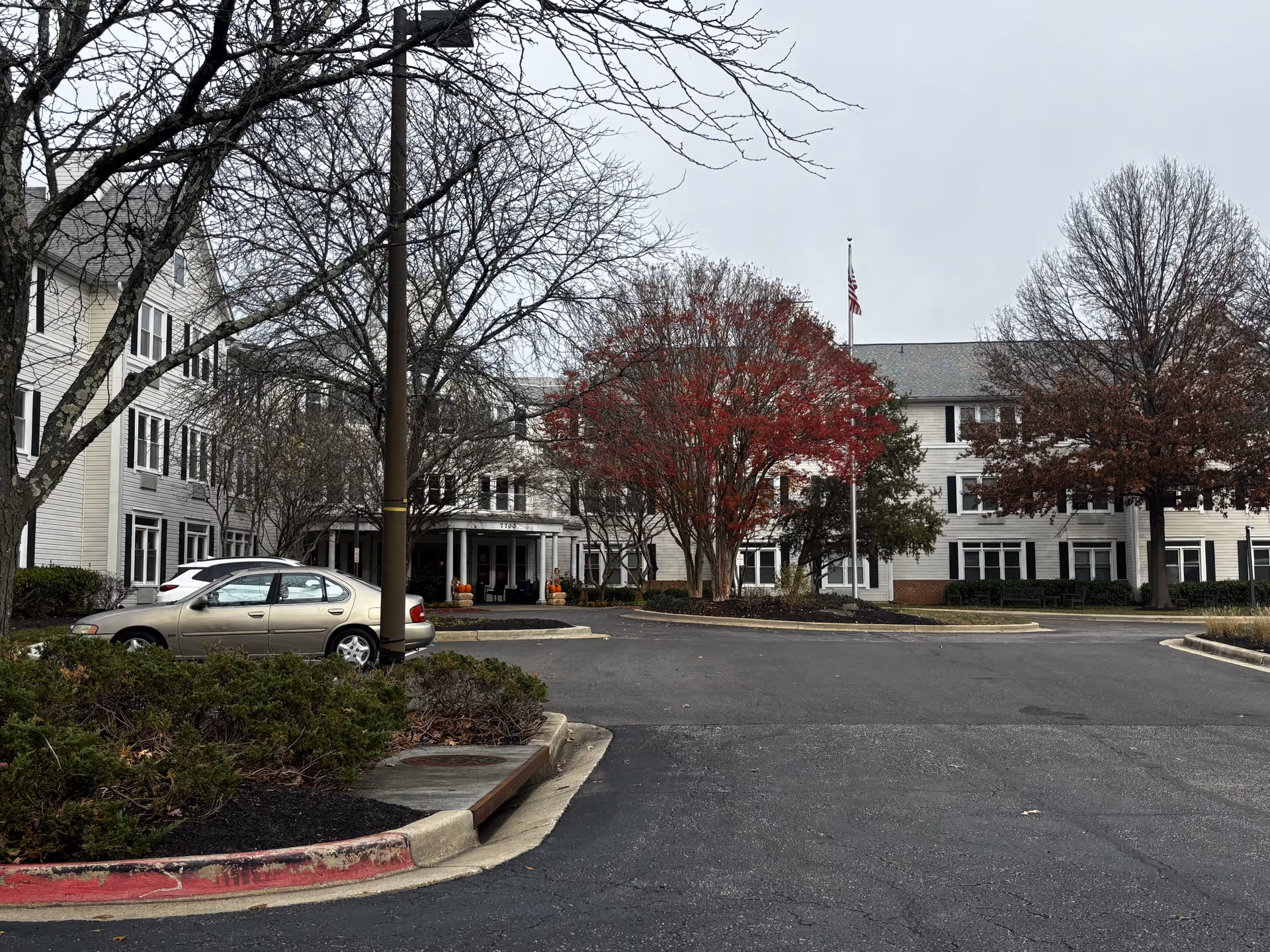 Exterior view of a large, three-story assisted living facility building with white siding and black shutters. There are several leafless trees and one tree with red leaves in front of the building. A flagpole with an American flag is visible near the center. There are a few parked cars and a driveway leading to the entrance, which is decorated with pumpkins.