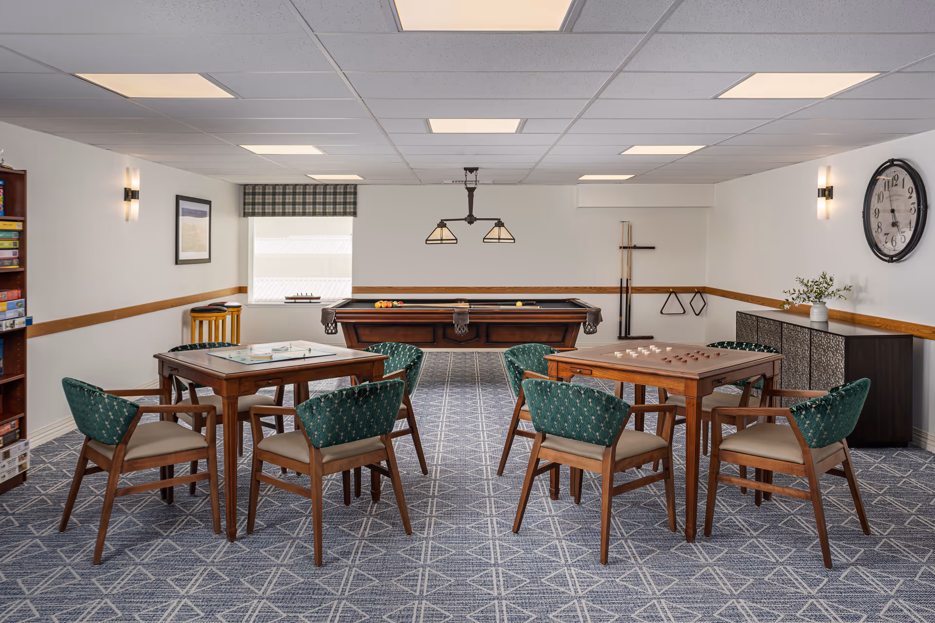 A recreational room in a senior living facility featuring two wooden tables with green cushioned chairs arranged around them. One table has a board game set up, and the other has a checkers game. In the background, there is a pool table with billiard balls and cues, a wall clock, and a cabinet with a vase and plant on top. The room has patterned carpet flooring, white walls, and ceiling lights.