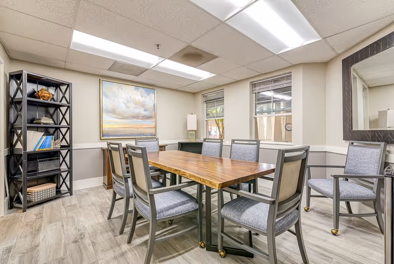 Well-lit dining/activity room featuring a rectangular wooden table surrounded by upholstered chairs, a bookshelf, wall art, and windows.