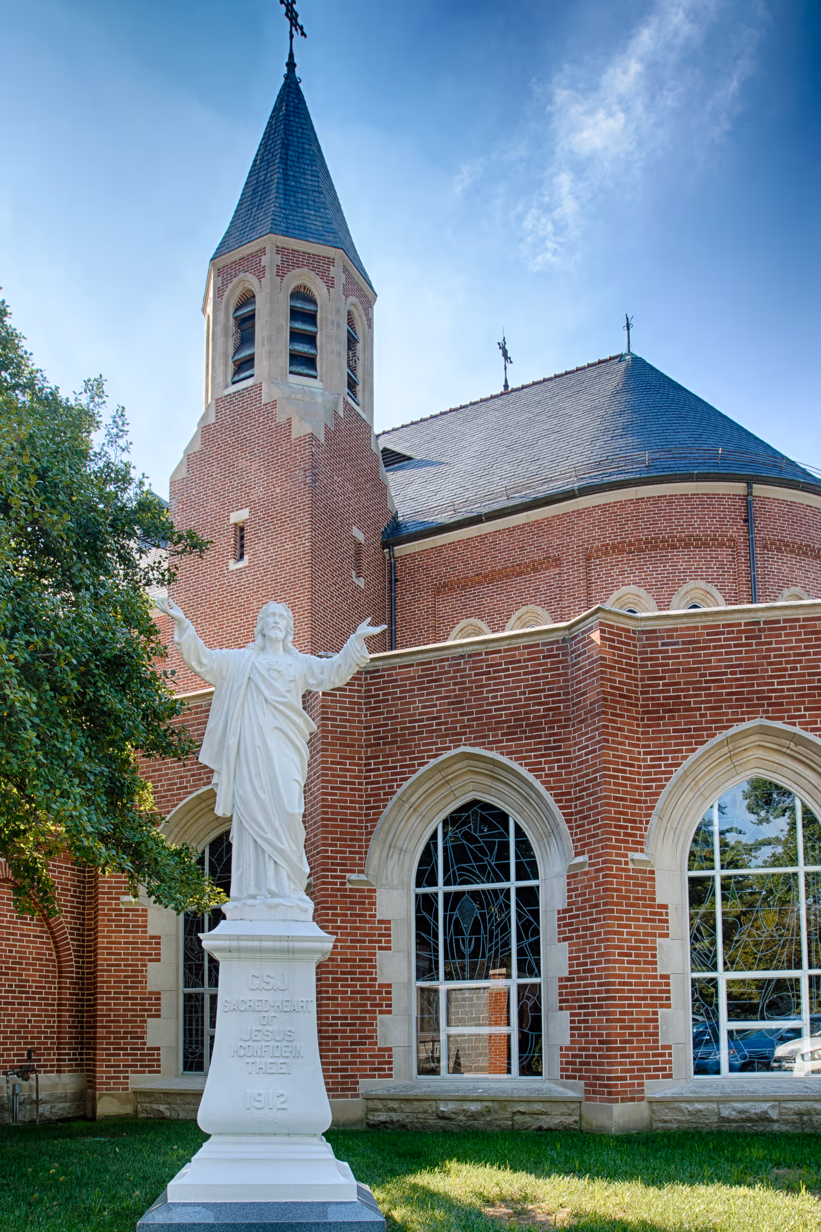 White statue of Jesus with outstretched arms on a pedestal stands on the lawn in front of a red-brick church with arched stained-glass windows and a steeple.