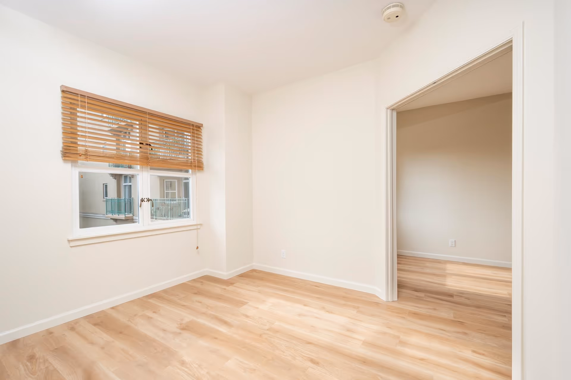 Empty room with light wood flooring, white walls, a window with wooden blinds, and an open doorway leading to another similar room.