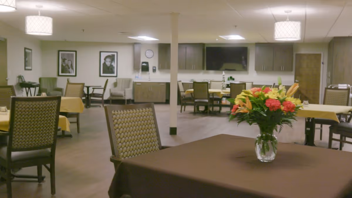 Community dining room with tables and chairs, a vase of flowers on a foreground table, and a kitchenette in the background.