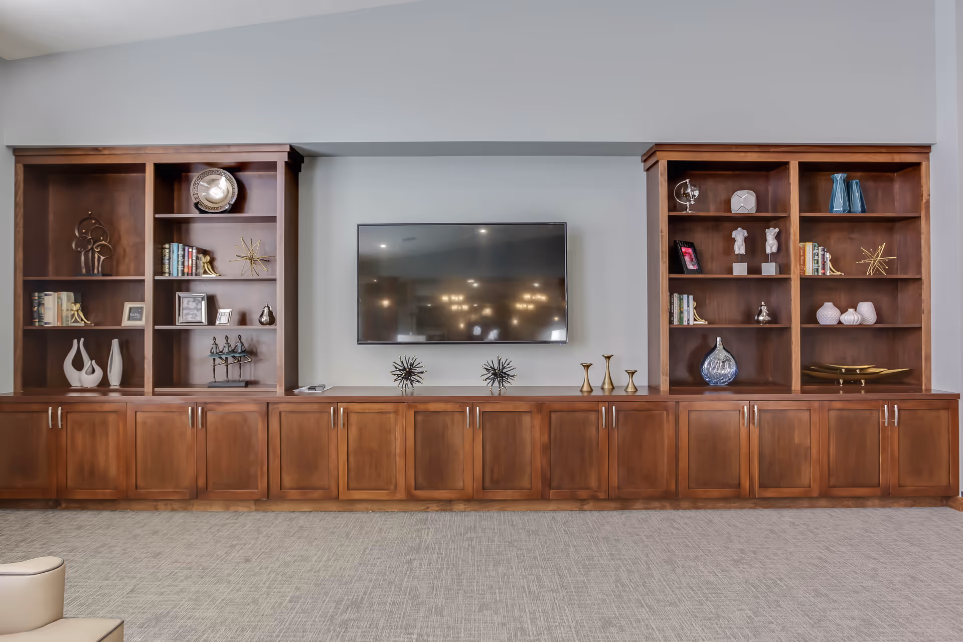 Wall-mounted TV centered between built-in wooden shelving and cabinets decorated with books and ornaments in a communal living area.