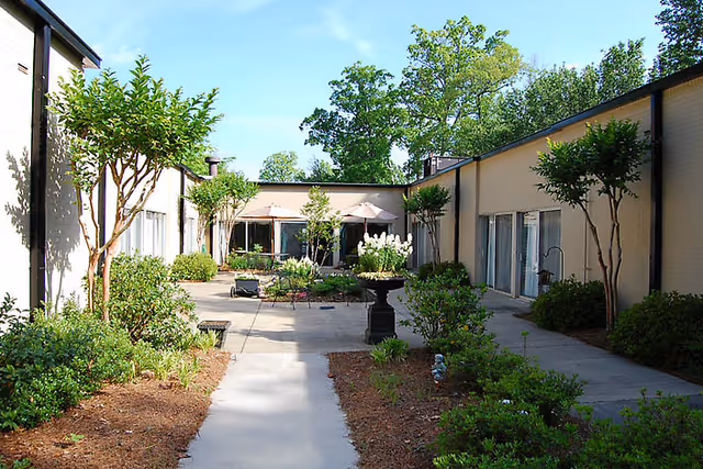 Sunlit central courtyard with a paved walkway, landscaped beds, small trees and patio umbrellas between single-story building wings.