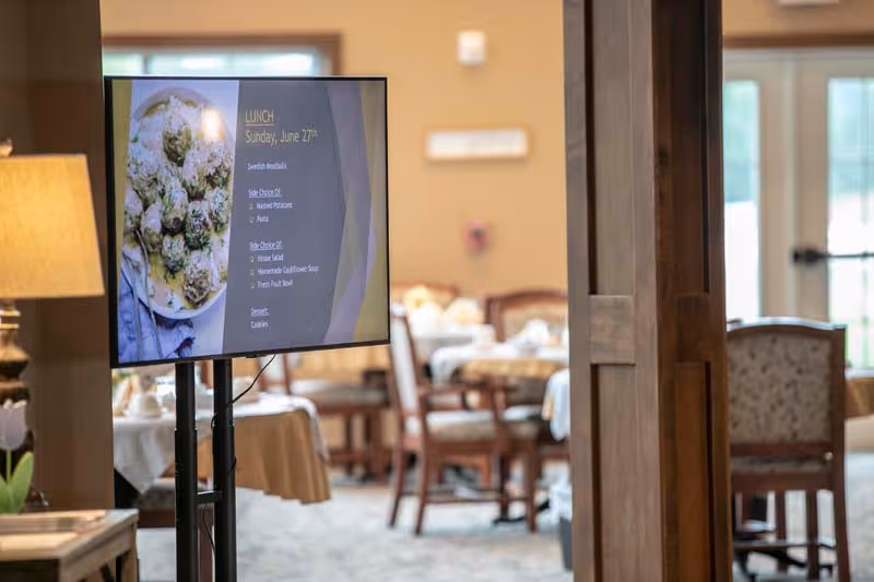 A digital screen displaying the lunch menu for Sunday, June 27th, placed in a dining room with tables and chairs set for a meal. The menu includes seafood meatballs, mashed potatoes, pasta, house salad, homemade cauliflower soup, fresh fruit bowl, and cookies. The room has warm lighting and large windows in the background.