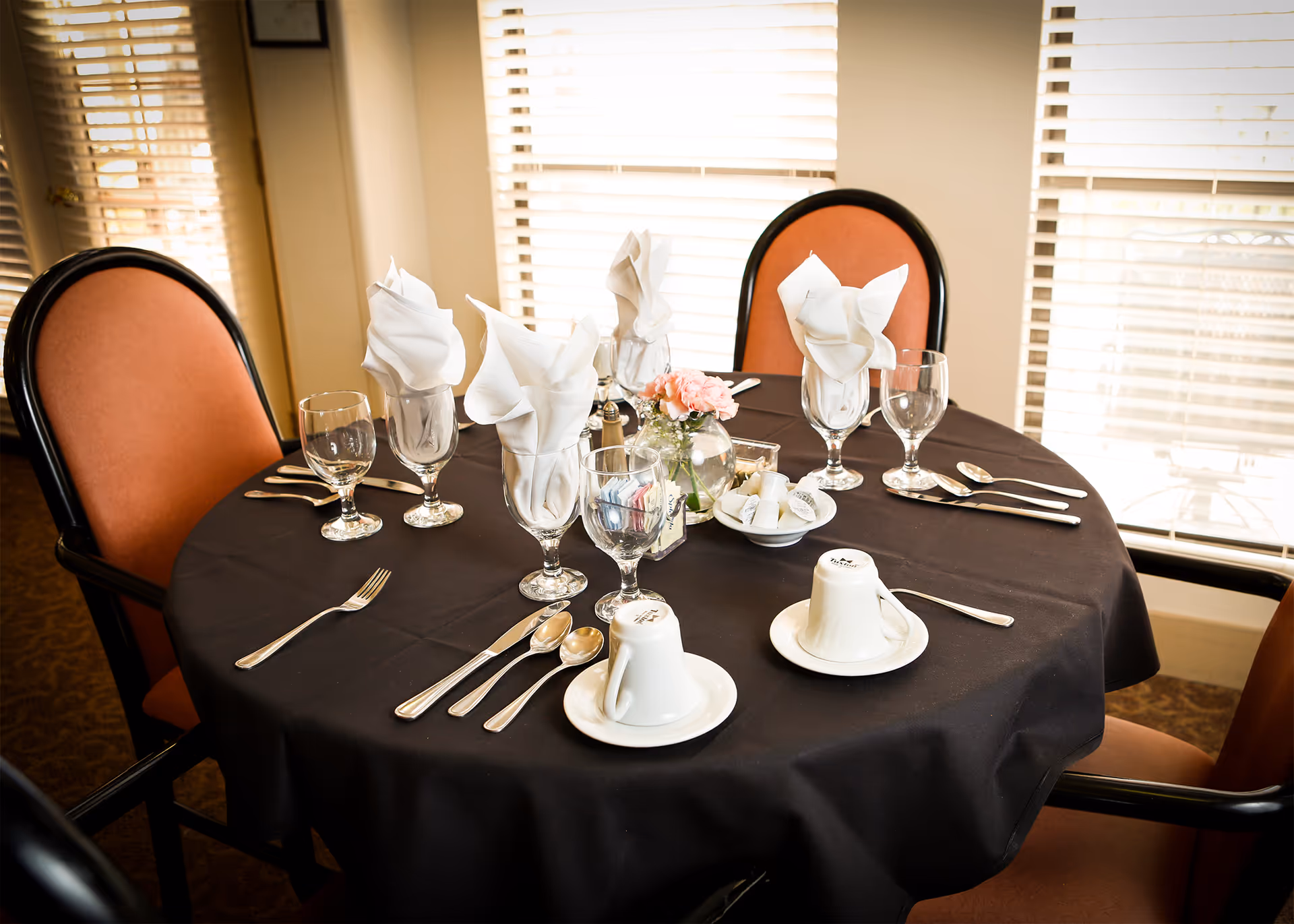 A round dining table set for four with a black tablecloth, white folded napkins in glasses, silverware, cups on saucers, and a small vase with pink flowers in the center. The table is surrounded by chairs with orange cushions and black frames, positioned near windows with white blinds.