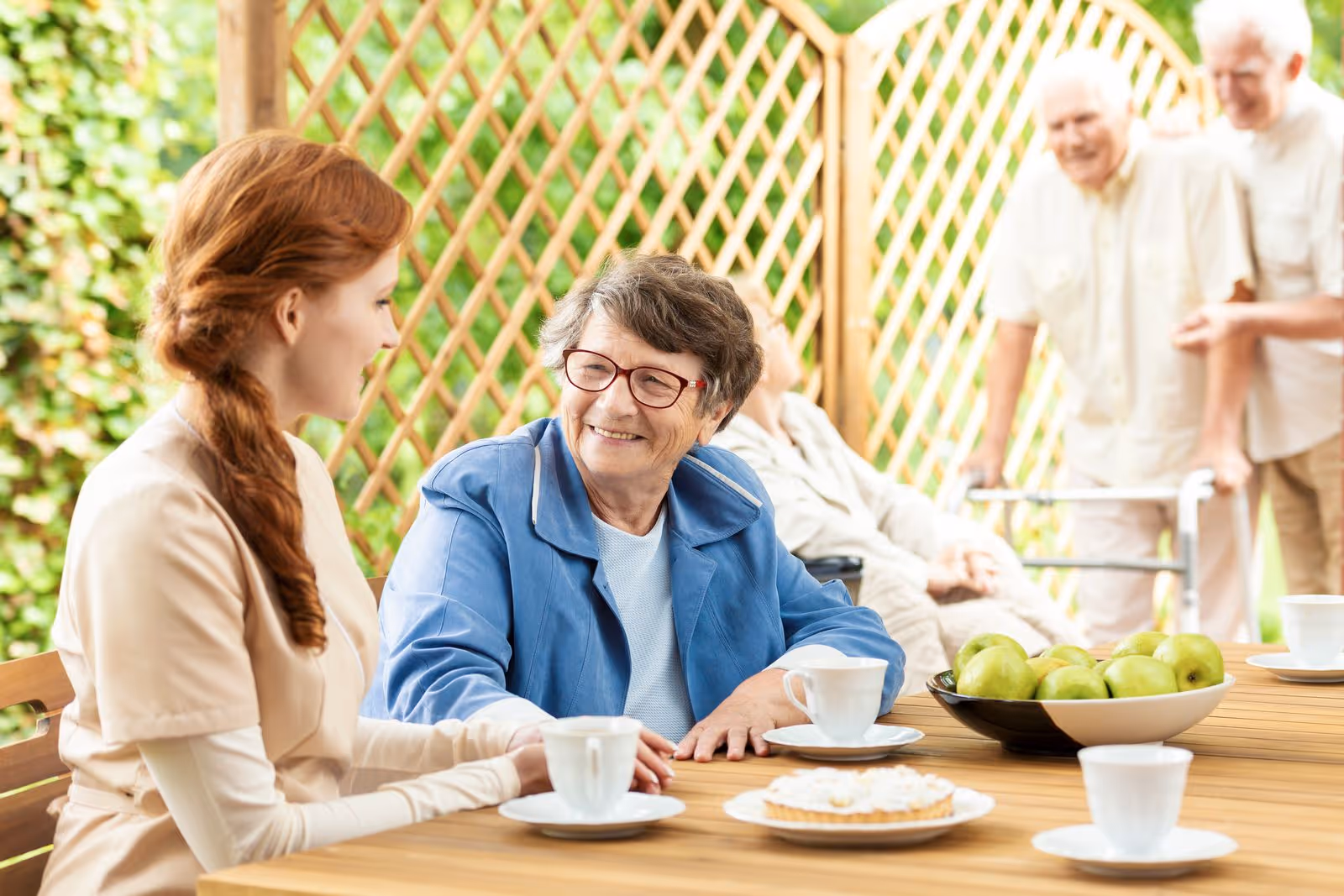 A young female caregiver with red hair talks and smiles with an elderly woman wearing glasses and a blue jacket while sitting at a wooden table outdoors. On the table are cups, a plate with a pastry, and a bowl of green apples. In the background, two elderly men, one using a walker, are also present near a wooden lattice fence with greenery.