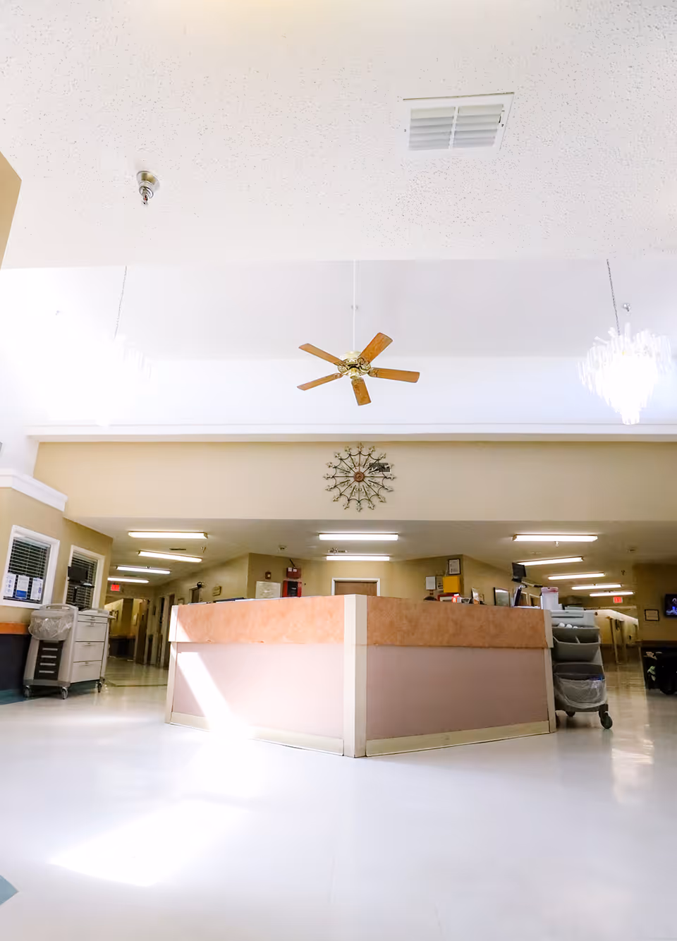 Bright senior living facility interior showing a central reception/nurse station with a ceiling fan and chandeliers.