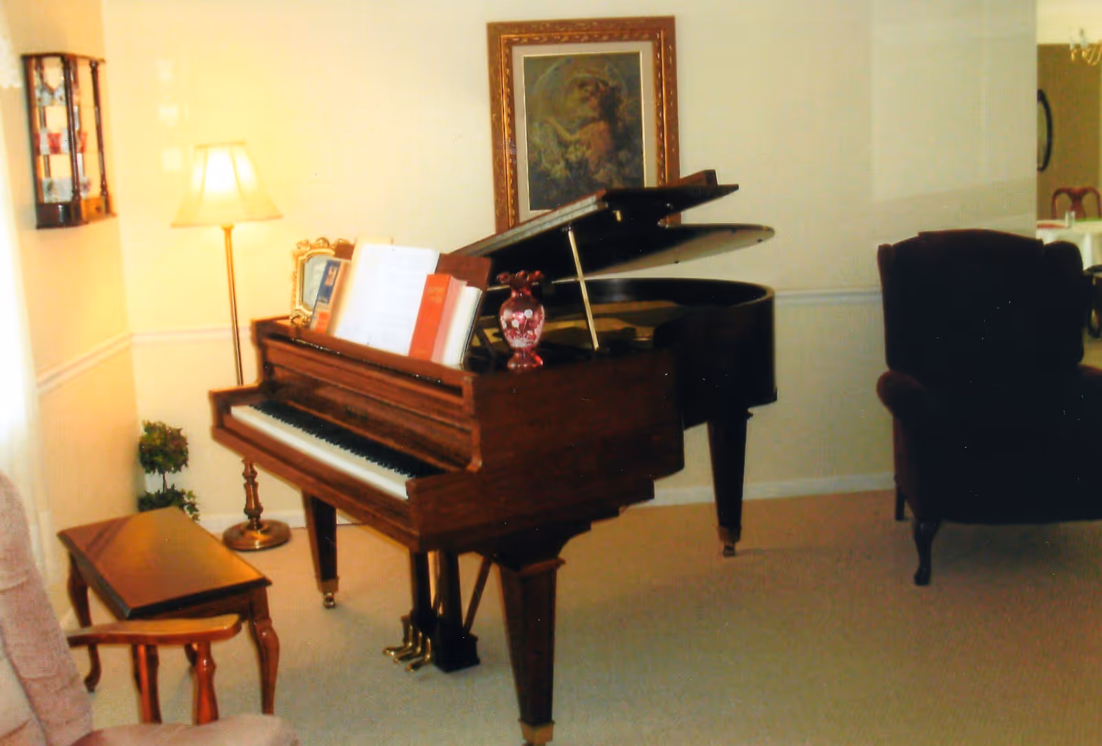 A cozy living room area featuring a grand piano with sheet music and a decorative vase on top. There is a floor lamp, a framed painting on the wall, a wooden bench, and an upholstered armchair. The room has light-colored walls and carpeted flooring.