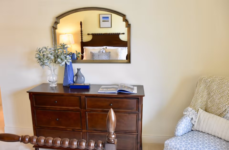 A cozy bedroom corner featuring a wooden dresser with decorative vases and an open book on top. Above the dresser is a large mirror reflecting a bed with pillows and a lamp. To the right, there is a comfortable armchair with a patterned blue and white cover and a beige throw blanket.