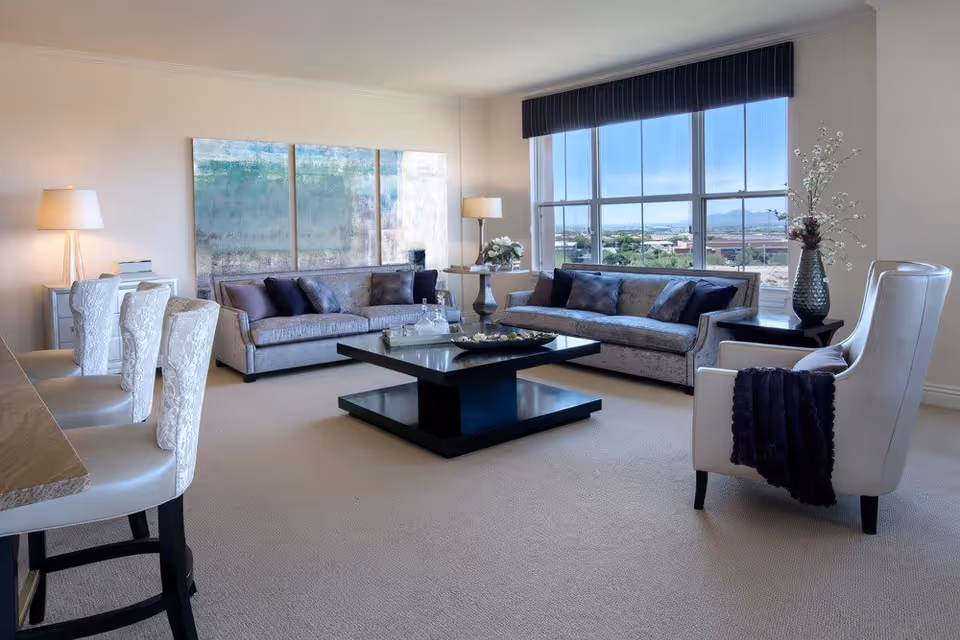 A bright and spacious living room with two gray sofas, a white armchair with a dark throw blanket, a large black coffee table, and two side tables with lamps and decorative items. Large windows with a view of the outdoors let in natural light, and a large abstract painting hangs on the wall behind one sofa.