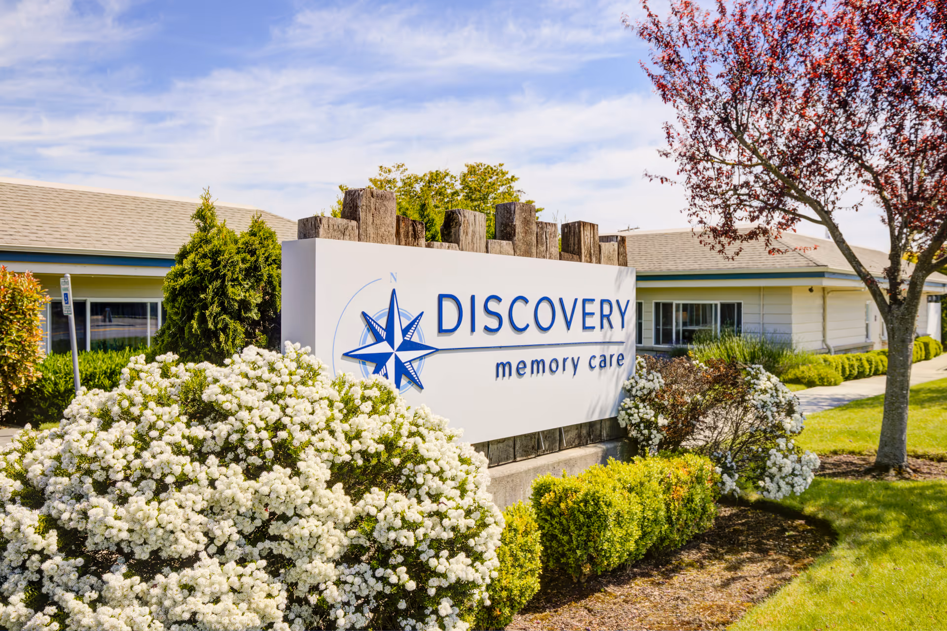 A landscaped entrance sign reading "DISCOVERY memory care" in front of a single-story building with flowering shrubs and a tree.