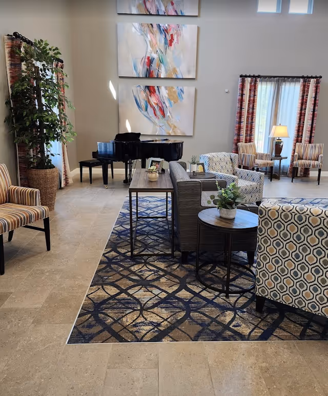 A spacious living room area with a grand piano in the corner, surrounded by various patterned armchairs and sofas arranged on a large decorative rug. The room features tall windows with colorful curtains, a side table with a lamp, and abstract artwork hanging on the wall above the piano. There is also a large potted plant near the window.