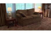 A cozy living room area featuring a brown loveseat with two cushions, flanked by two wooden side tables each holding a lamp. The room has large windows with sheer white curtains and heavier drapes, and a patterned carpet covering the floor.