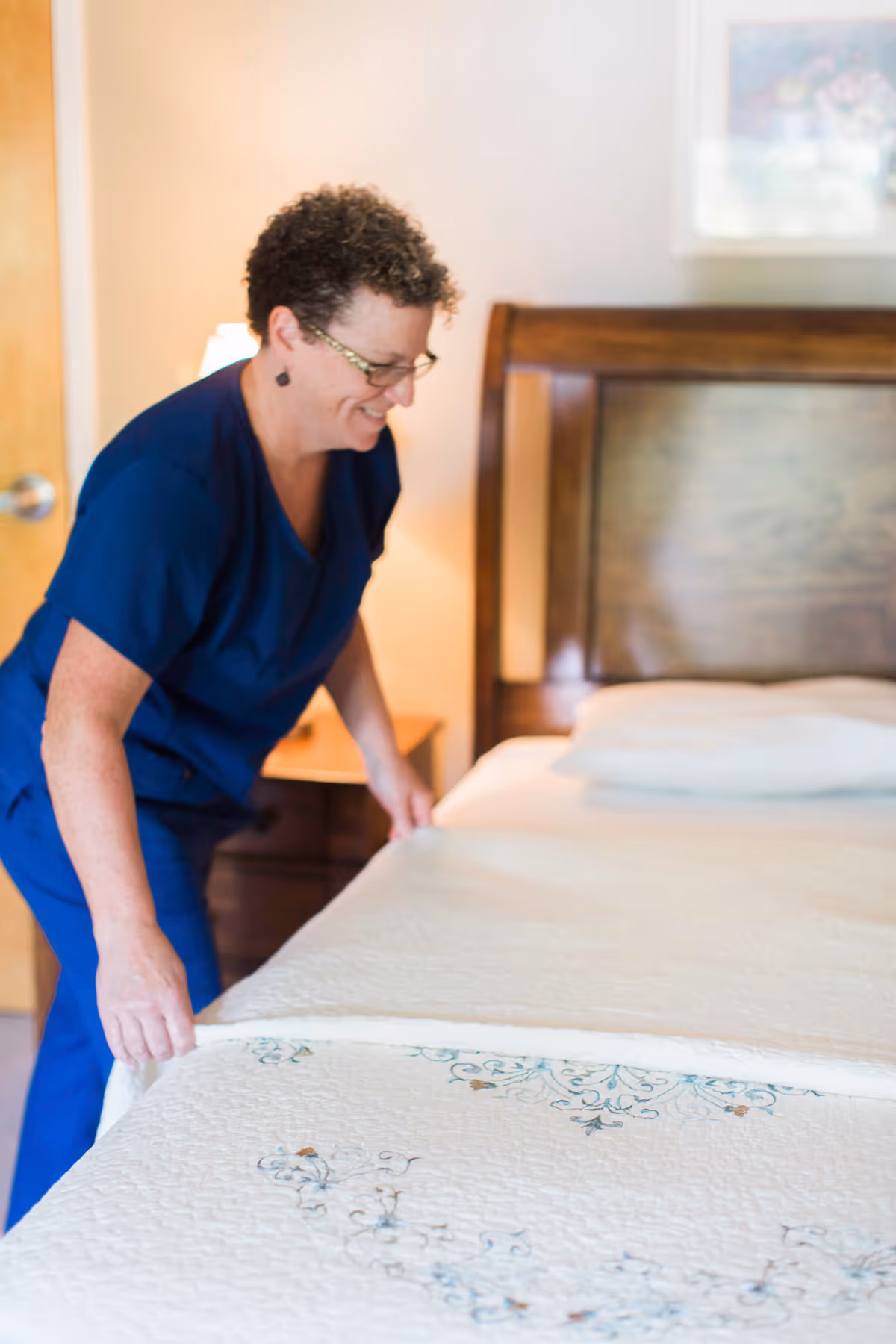 A woman in navy scrubs is making a bed in a bedroom with a wooden headboard.