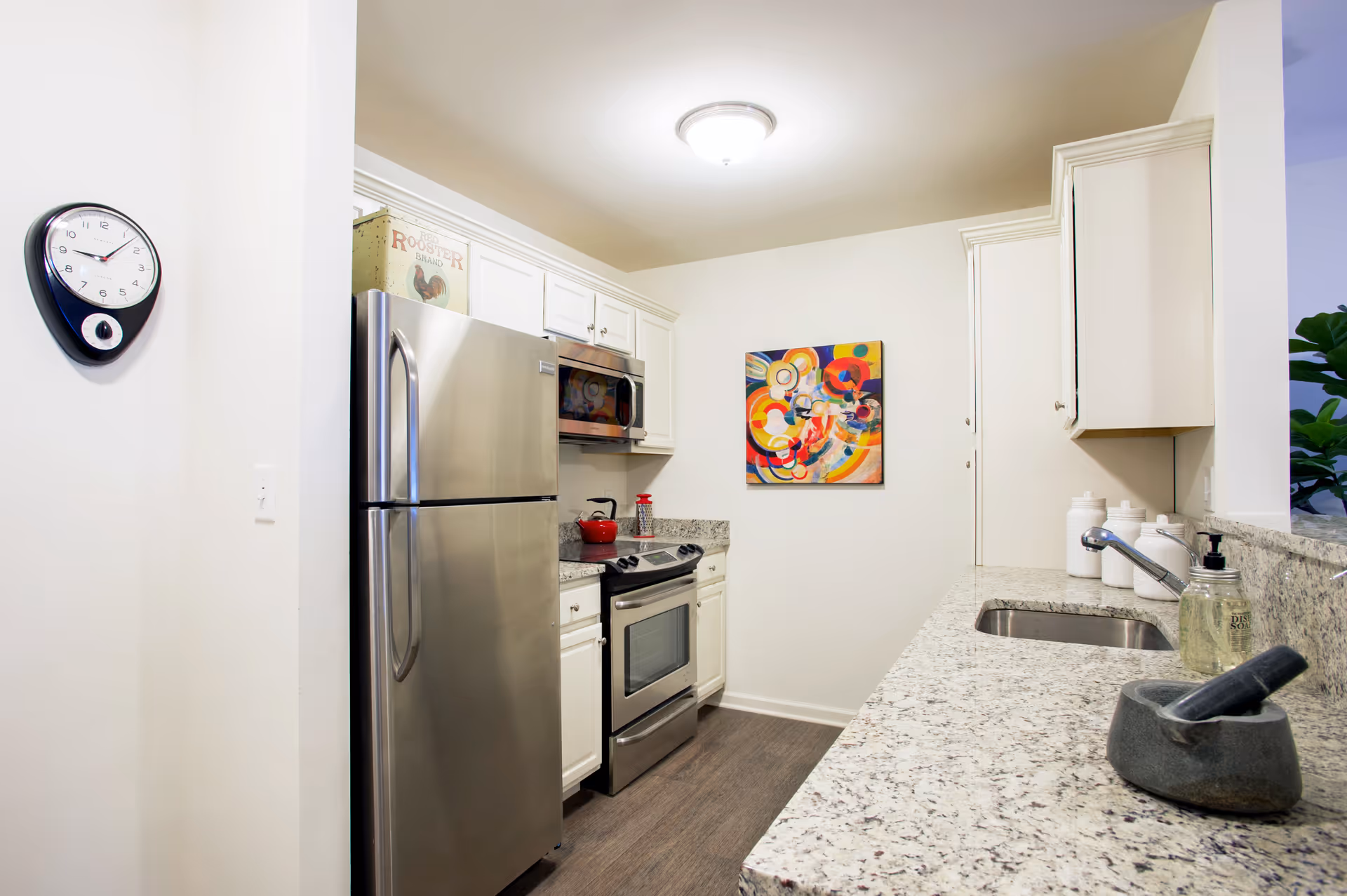 A modern kitchen with white cabinets, a stainless steel refrigerator, stove, and microwave. The kitchen features granite countertops with a sink, a mortar and pestle, and a soap dispenser. A colorful abstract painting hangs on the wall, and a clock is mounted on a nearby wall.
