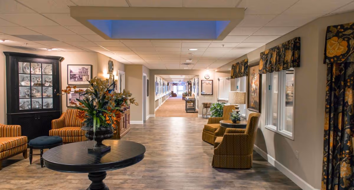 A well-lit hallway in a senior living facility with a round table holding a floral arrangement in the foreground. The hallway features comfortable seating areas with armchairs and side tables, decorative curtains on the windows, framed pictures on the walls, and a wooden floor. The corridor extends into the distance with additional furniture and lighting.