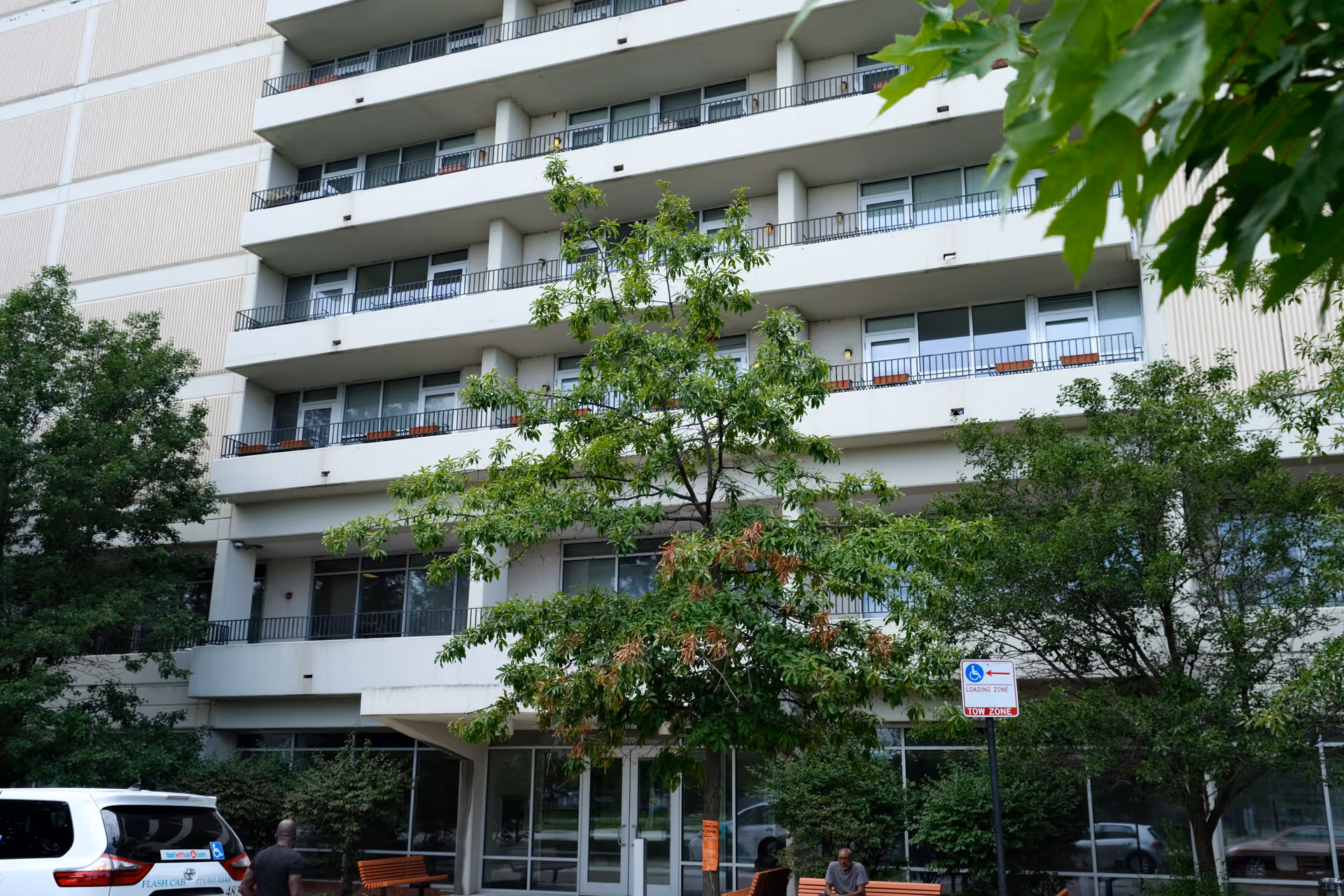 Front facade of a multi-story residential building with balconies, trees, benches and an entrance.