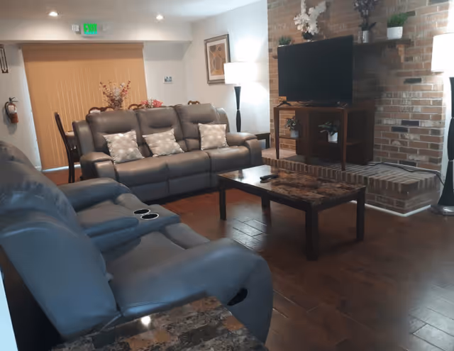 A cozy living room with two gray leather sofas, a coffee table with a marble-like surface, a TV on a wooden stand in front of a brick fireplace, and a floor lamp. There are decorative pillows on the sofas, a framed picture on the wall, and a dining area visible in the background with a table and chairs.